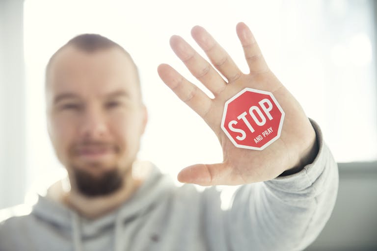A man in a hoodie holds up his hand with a 'Stop and Pray' sticker. Focus on hand and sticker.
