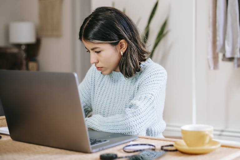 Focused young woman using laptop at home while drinking coffee. Ideal for remote work and study themes.